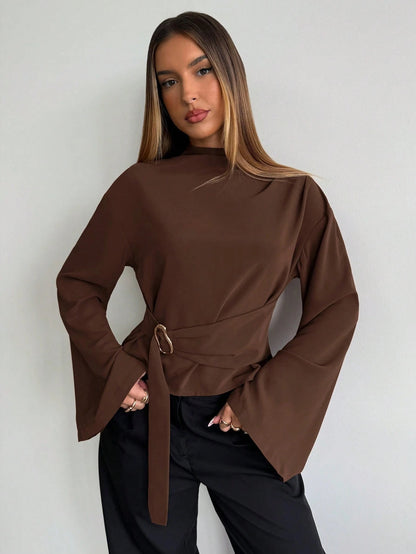 Woman wearing a coffee brown mock-neck long-sleeve top with bell sleeves and an integrated waist belt with a gold buckle against a white wall.