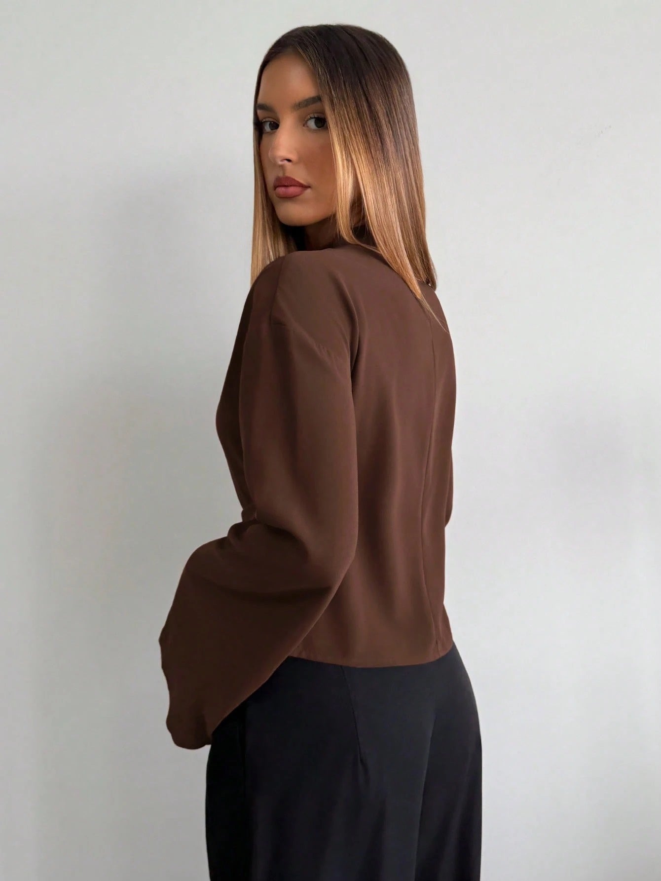 Woman wearing a coffee brown mock-neck long-sleeve top with bell sleeves and an integrated waist belt with a gold buckle against a white wall.