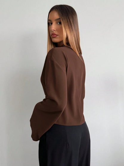 Woman wearing a coffee brown mock-neck long-sleeve top with bell sleeves and an integrated waist belt with a gold buckle against a white wall.