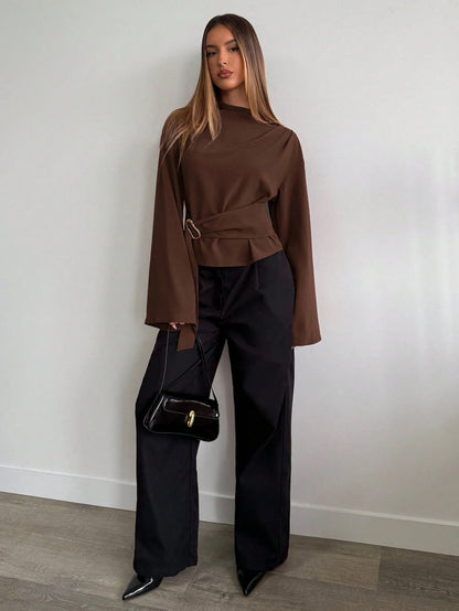 Woman wearing a coffee brown mock-neck long-sleeve top with bell sleeves and an integrated waist belt with a gold buckle against a white wall.