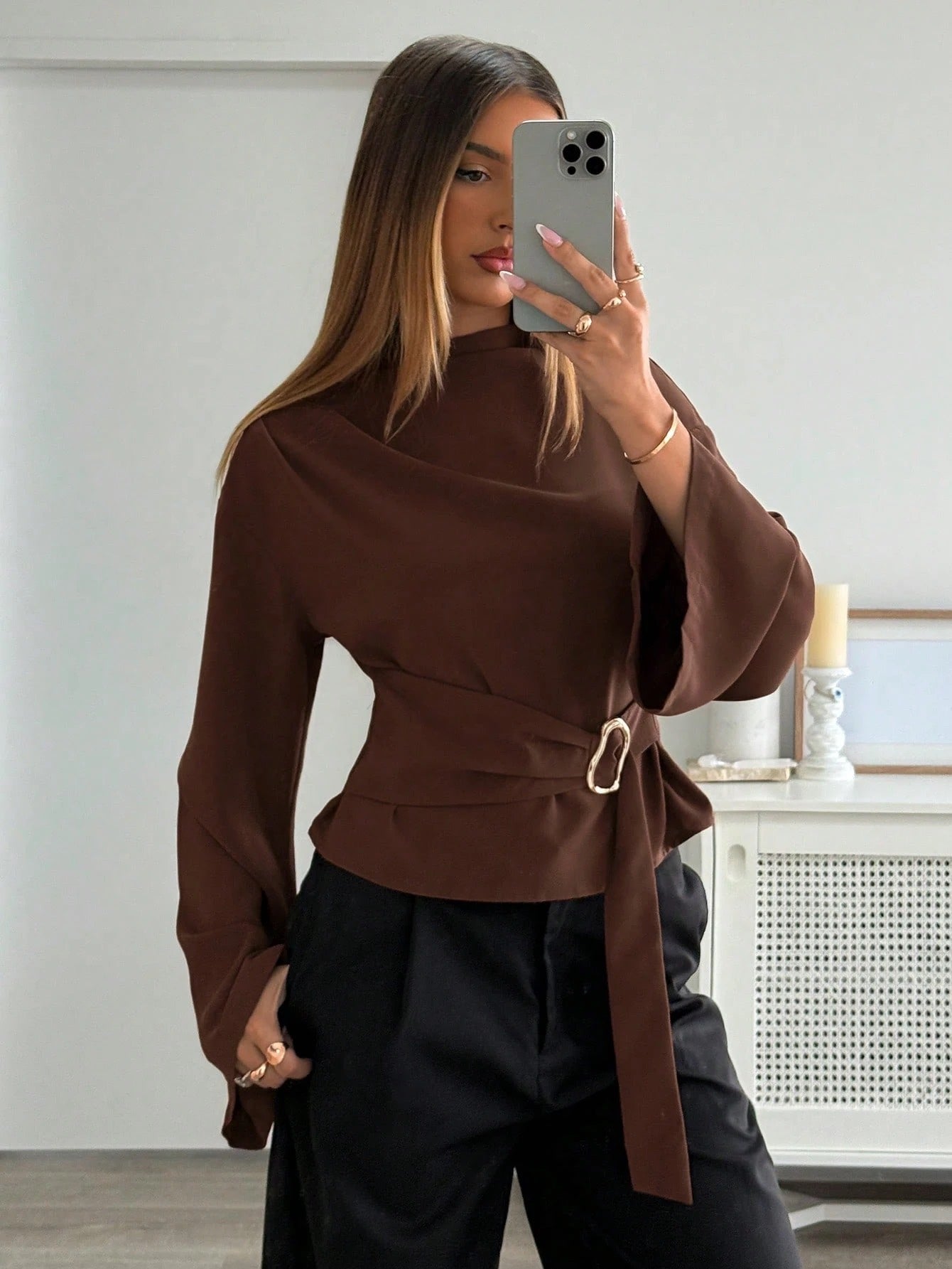 Woman wearing a coffee brown mock-neck long-sleeve top with bell sleeves and an integrated waist belt with a gold buckle against a white wall.