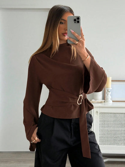 Woman wearing a coffee brown mock-neck long-sleeve top with bell sleeves and an integrated waist belt with a gold buckle against a white wall.
