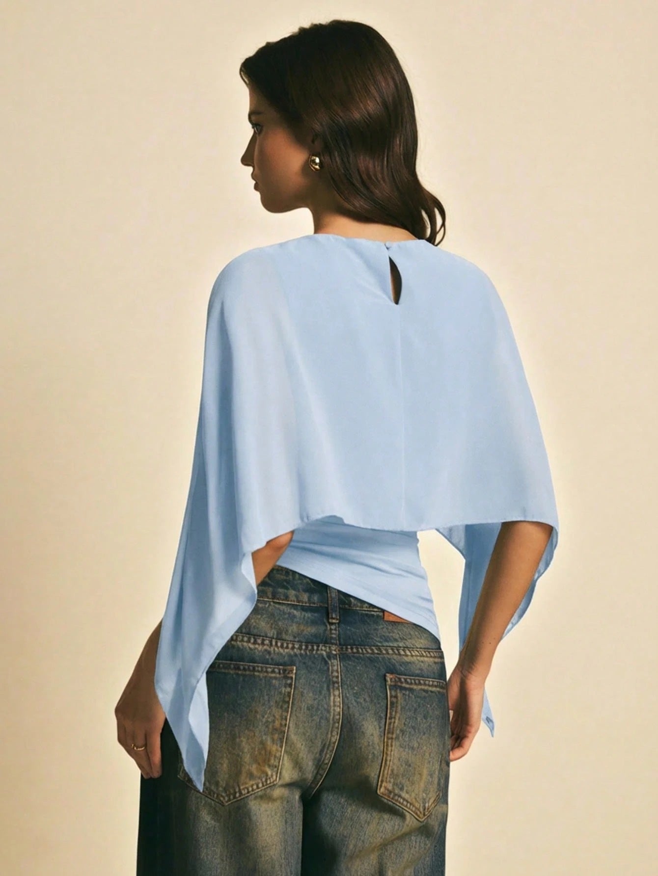 Woman wearing a light blue solid color elegant shawl blouse with cape sleeves paired with dark denim jeans