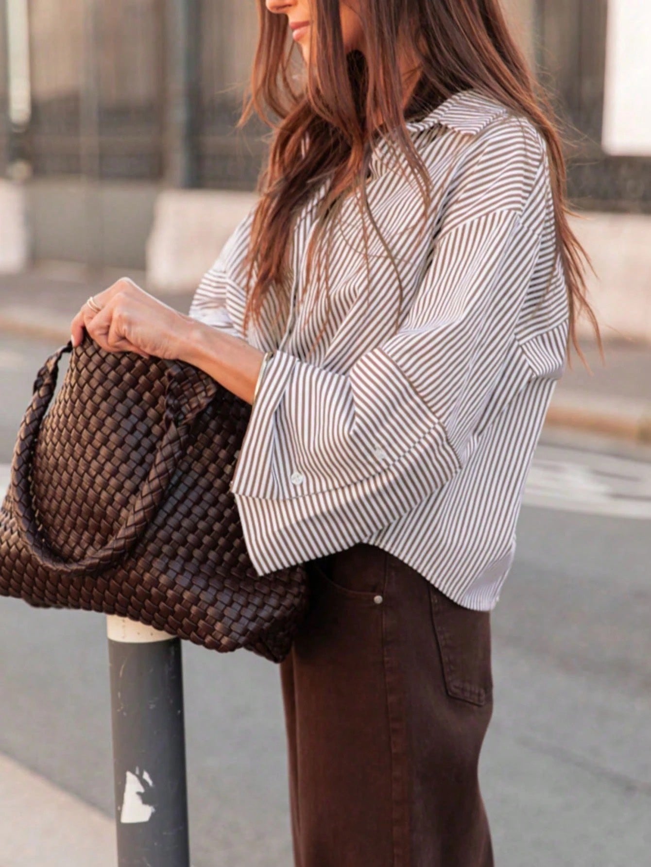 Woman wearing a light khaki vertical striped drop-shoulder button-down shirt paired with brown pants, holding a dark woven handbag