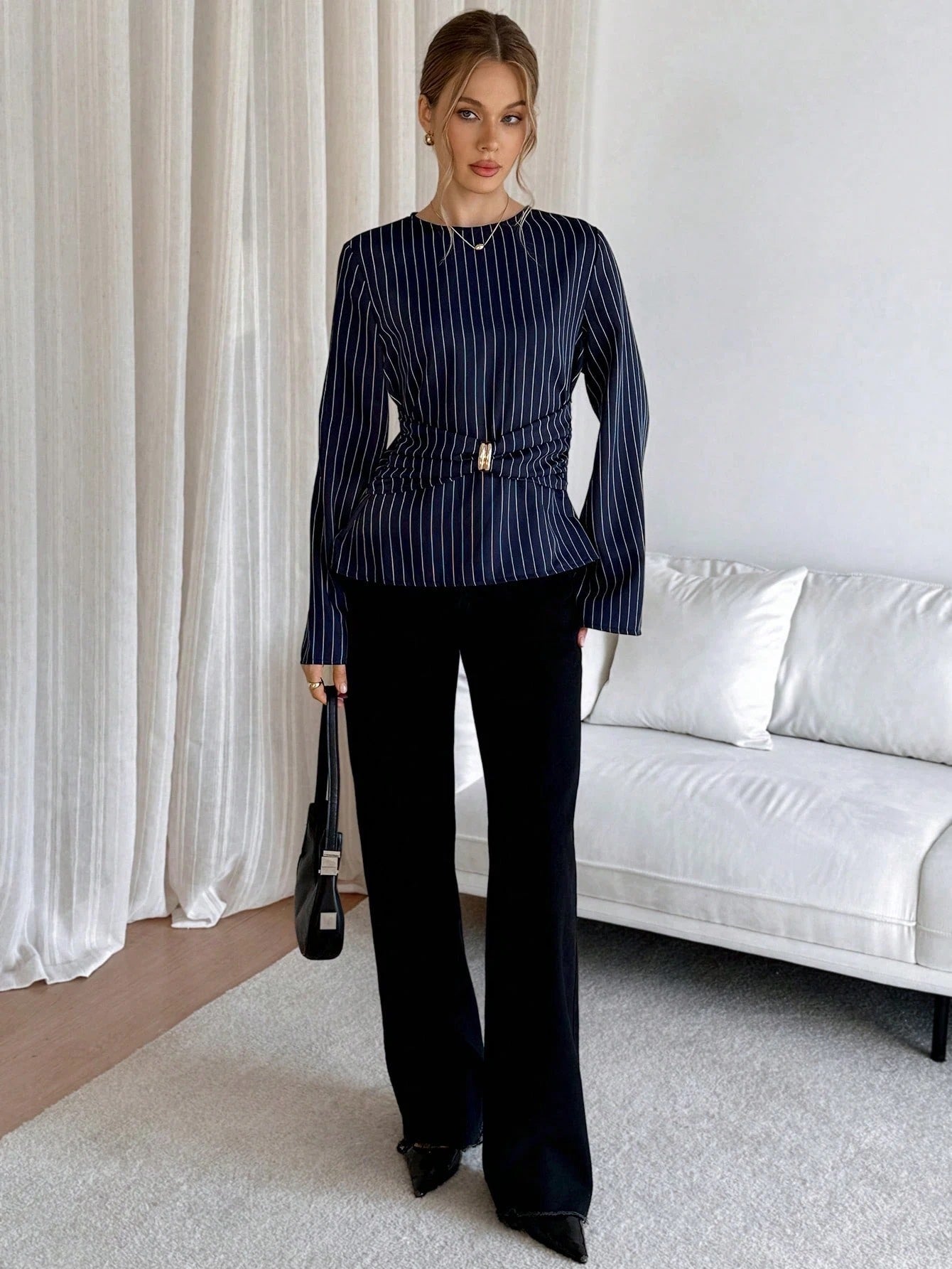 Woman wearing a navy blue pinstripe long sleeve blouse featuring a metal ring decor waist detail and dark trousers