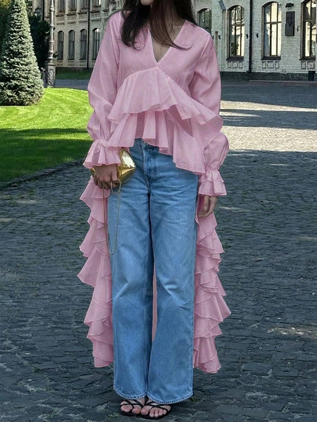 Woman wearing a pink long-sleeve V-neck top with an asymmetrical cascading ruffle high-low hem paired with blue jeans and holding a gold clutch.