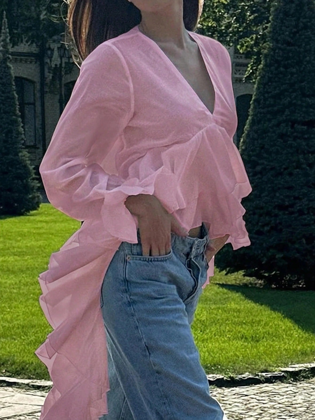 Woman wearing a pink long-sleeve V-neck top with an asymmetrical cascading ruffle high-low hem paired with blue jeans and holding a gold clutch.