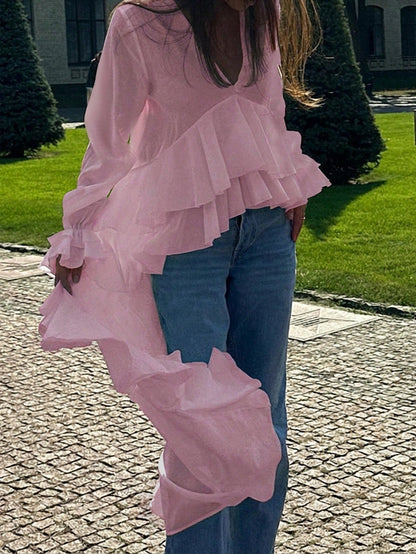 Woman wearing a pink long-sleeve V-neck top with an asymmetrical cascading ruffle high-low hem paired with blue jeans and holding a gold clutch.