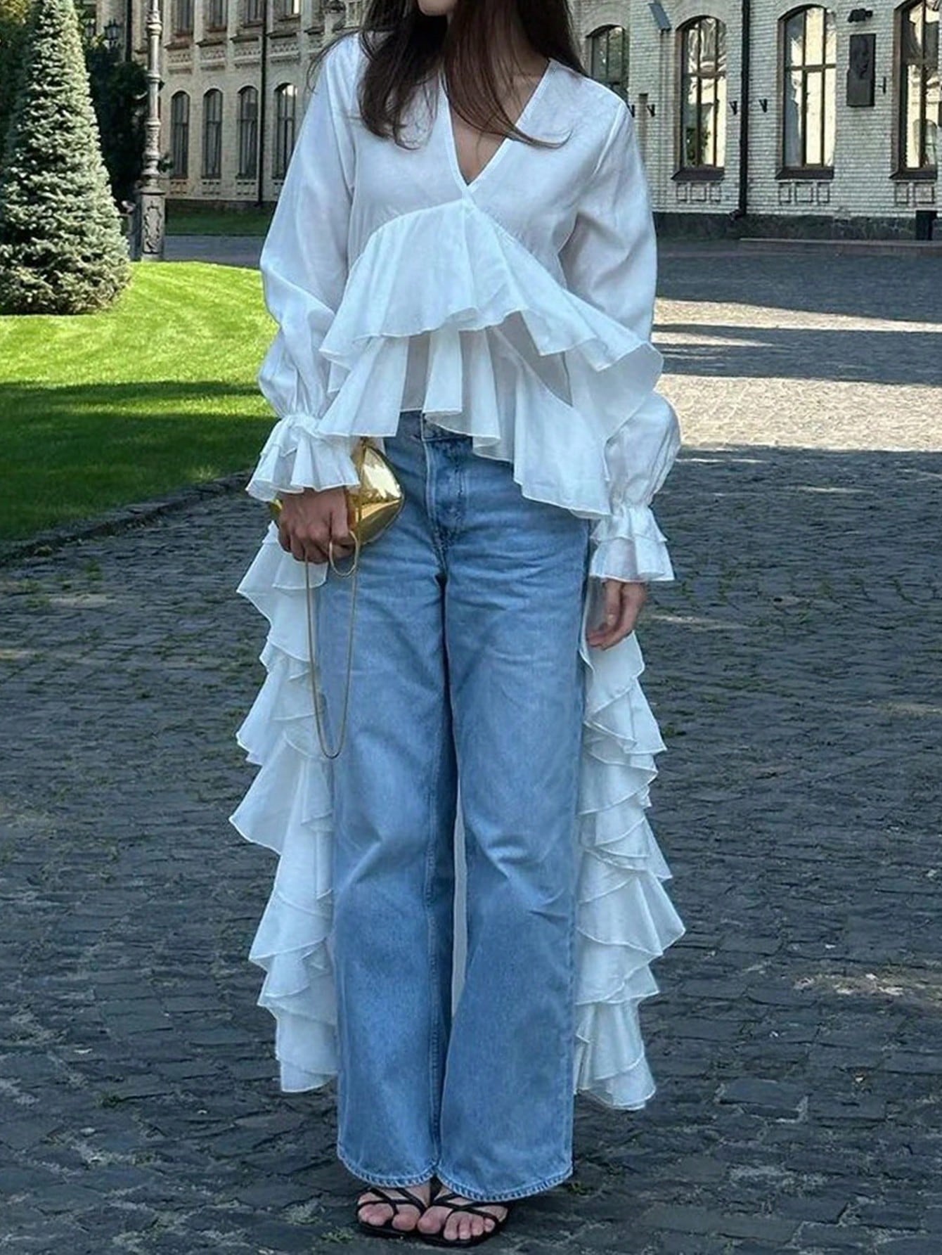 Woman wearing a white long-sleeve V-neck top with an asymmetrical cascading ruffle high-low hem paired with blue jeans and holding a gold clutch.