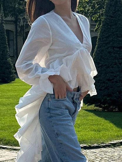 Woman wearing a white long-sleeve V-neck top with an asymmetrical cascading ruffle high-low hem paired with blue jeans and holding a gold clutch.