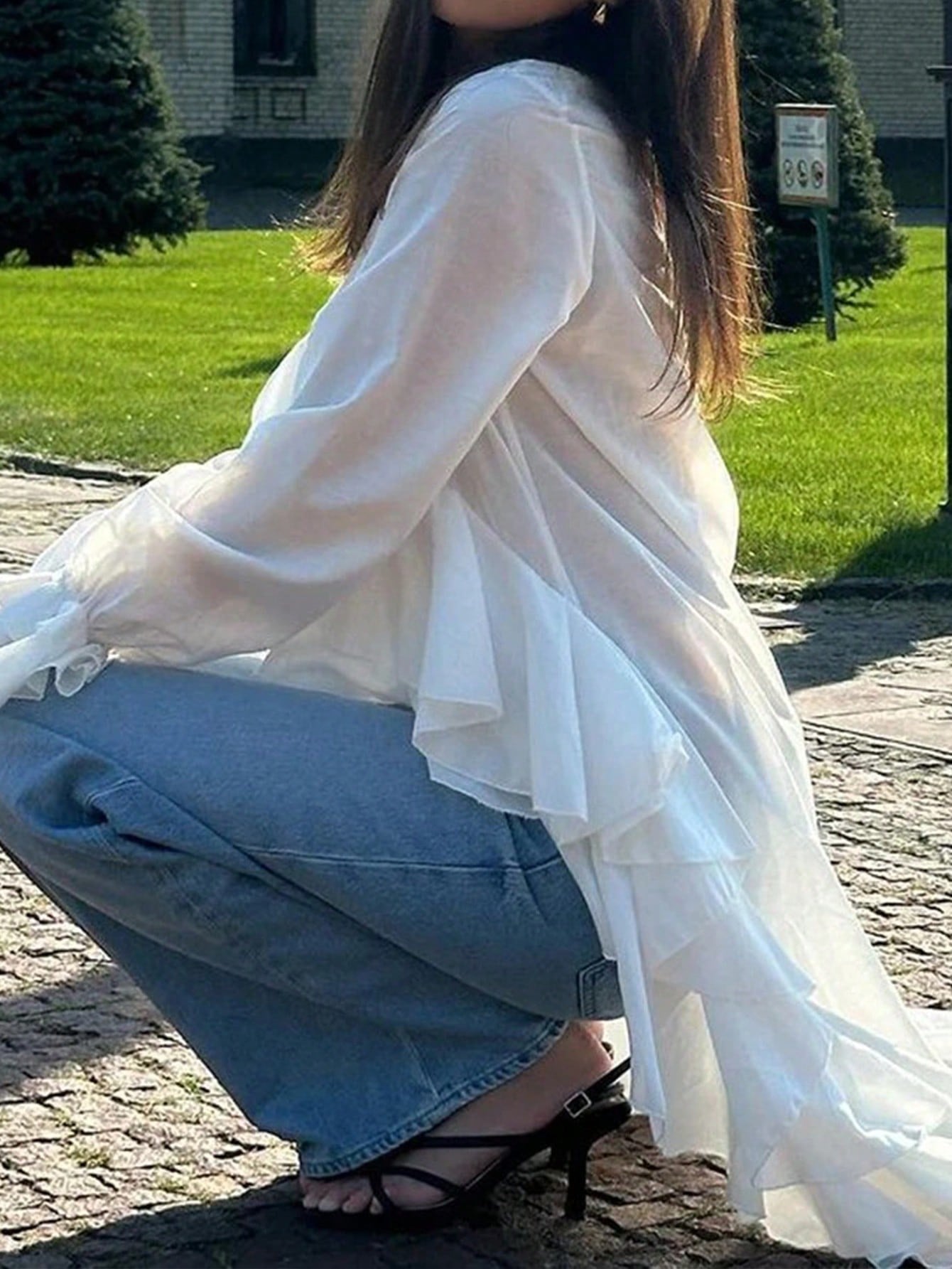 Woman wearing a white long-sleeve V-neck top with an asymmetrical cascading ruffle high-low hem paired with blue jeans and holding a gold clutch.