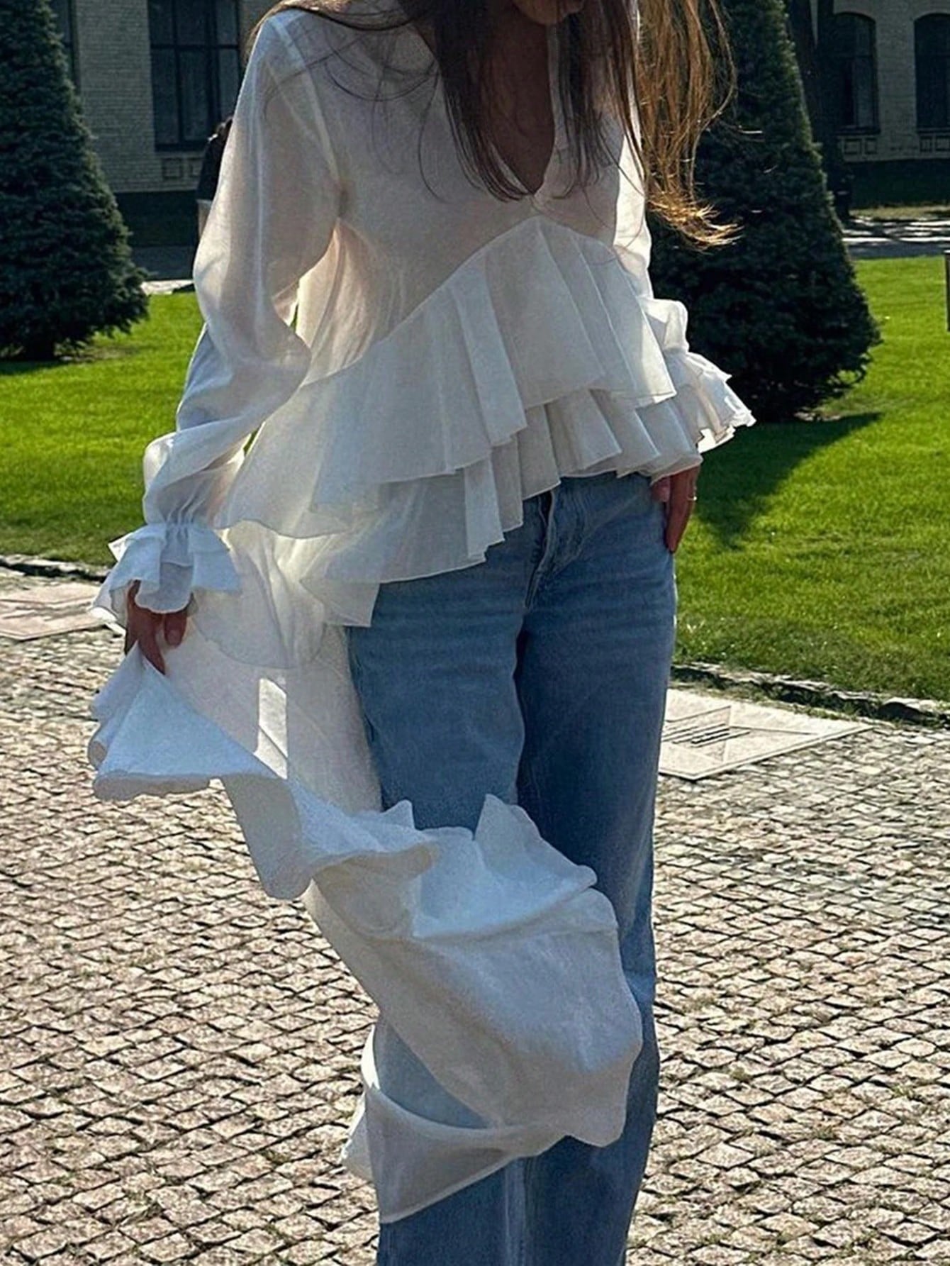 Woman wearing a white long-sleeve V-neck top with an asymmetrical cascading ruffle high-low hem paired with blue jeans and holding a gold clutch.