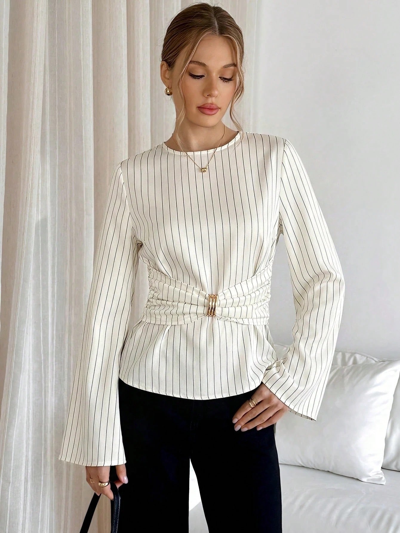 Woman wearing a white pinstripe long sleeve blouse featuring a metal ring decor waist detail and dark trousers