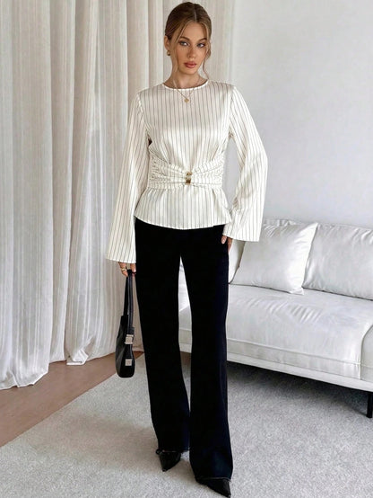 Woman wearing a white pinstripe long sleeve blouse featuring a metal ring decor waist detail and dark trousers
