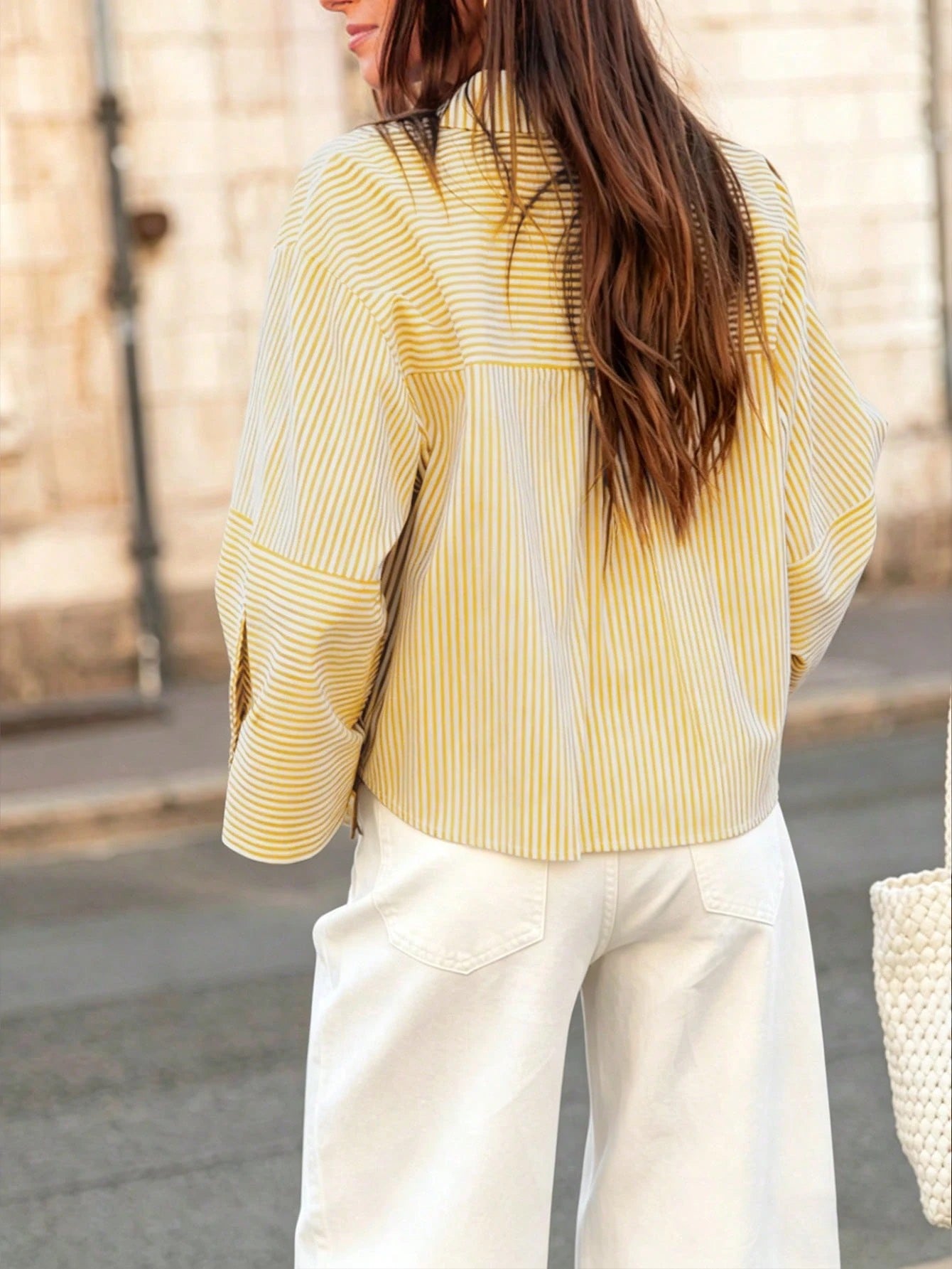 Woman wearing a yellow vertical striped drop-shoulder button-down shirt paired with white pants, holding a dark woven handbag