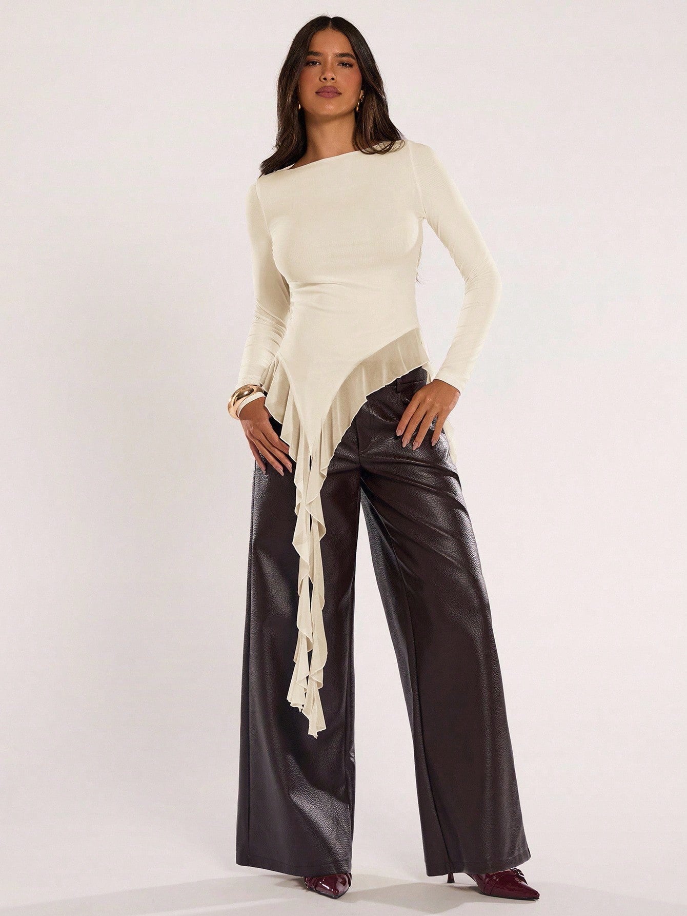Woman wearing an apricot fitted long sleeve top with an asymmetric ruffle hem paired with dark brown faux leather pants