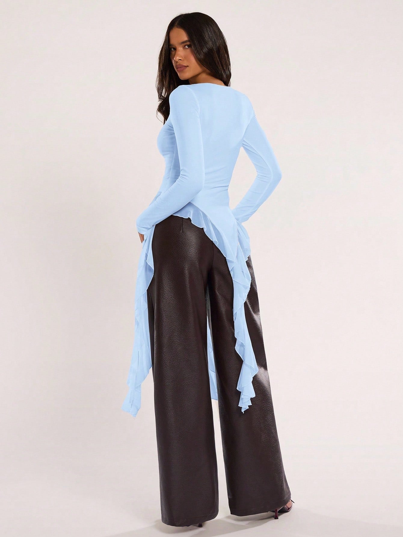 Woman wearing an baby blue fitted long sleeve top with an asymmetric ruffle hem paired with dark brown faux leather pants