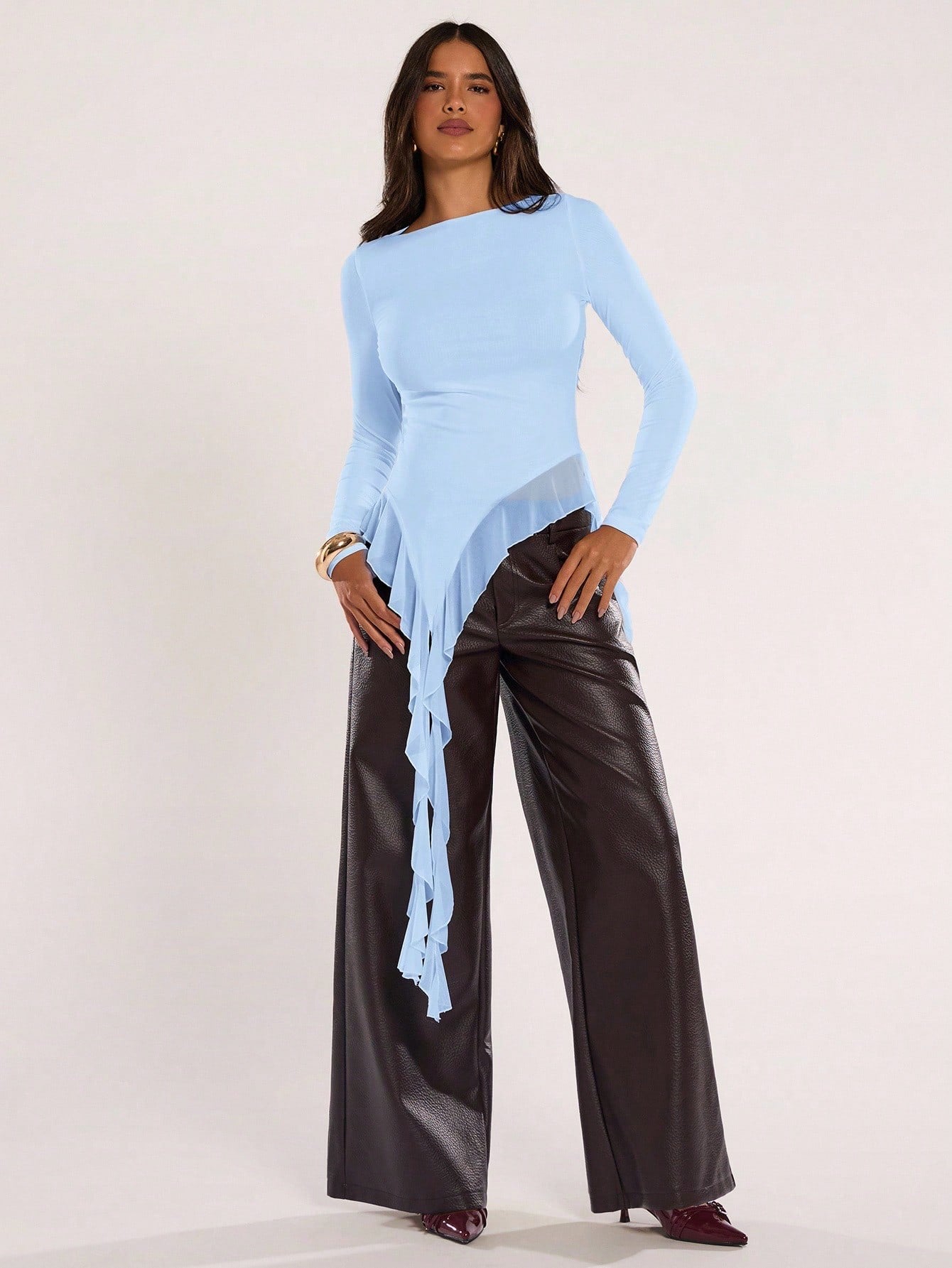 Woman wearing an baby blue fitted long sleeve top with an asymmetric ruffle hem paired with dark brown faux leather pants