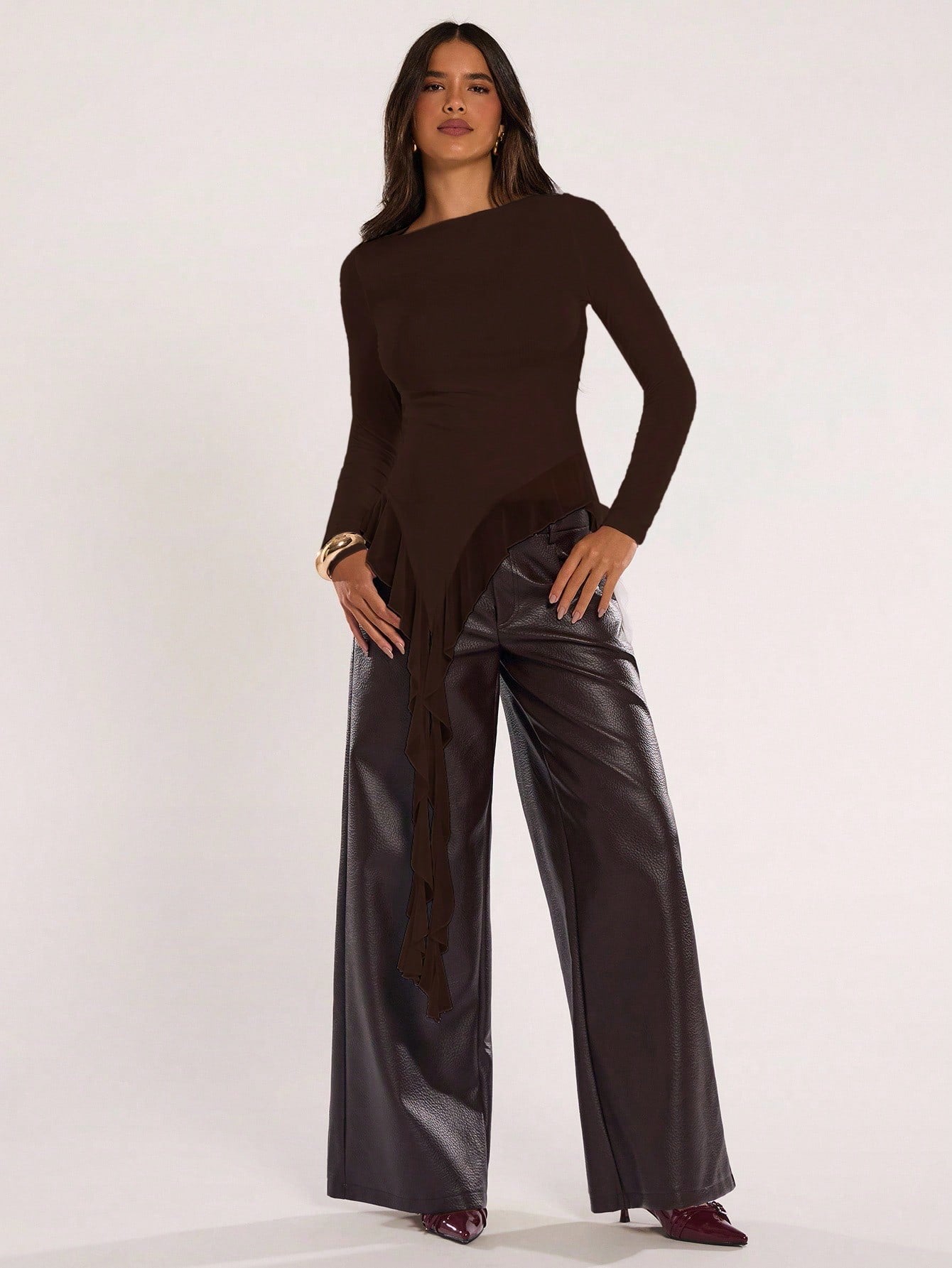 Woman wearing an coffee brown fitted long sleeve top with an asymmetric ruffle hem paired with dark brown faux leather pants