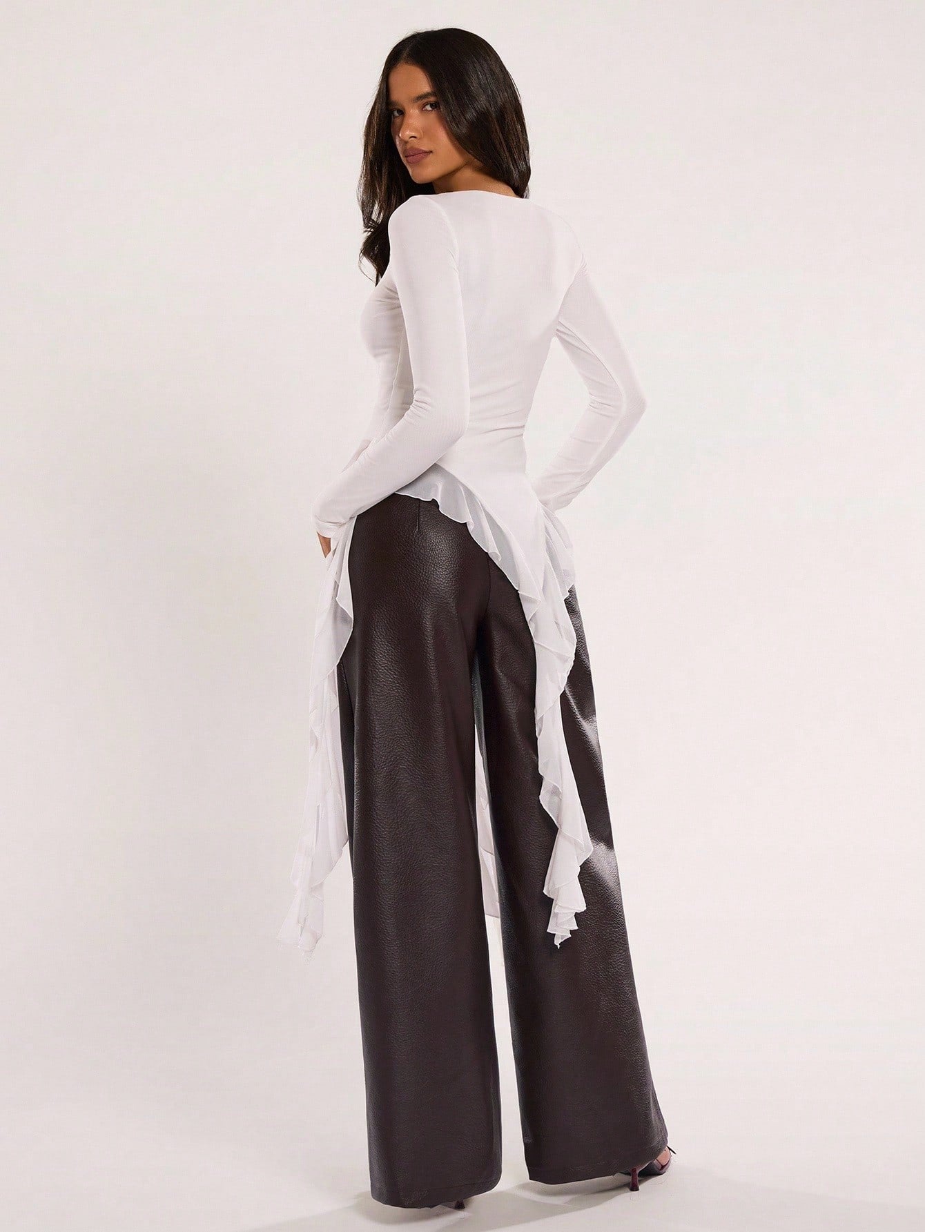 Woman wearing an off-white fitted long sleeve top with an asymmetric ruffle hem paired with dark brown faux leather pants