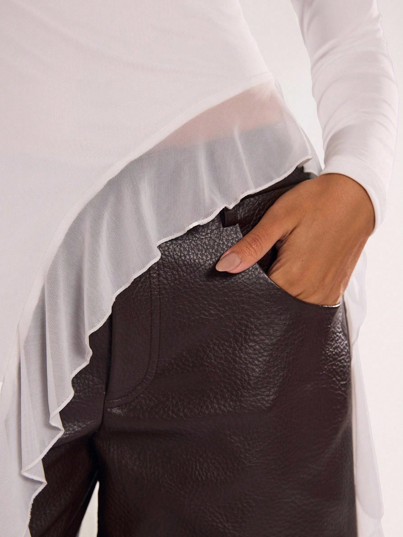 Woman wearing an off-white fitted long sleeve top with an asymmetric ruffle hem paired with dark brown faux leather pants