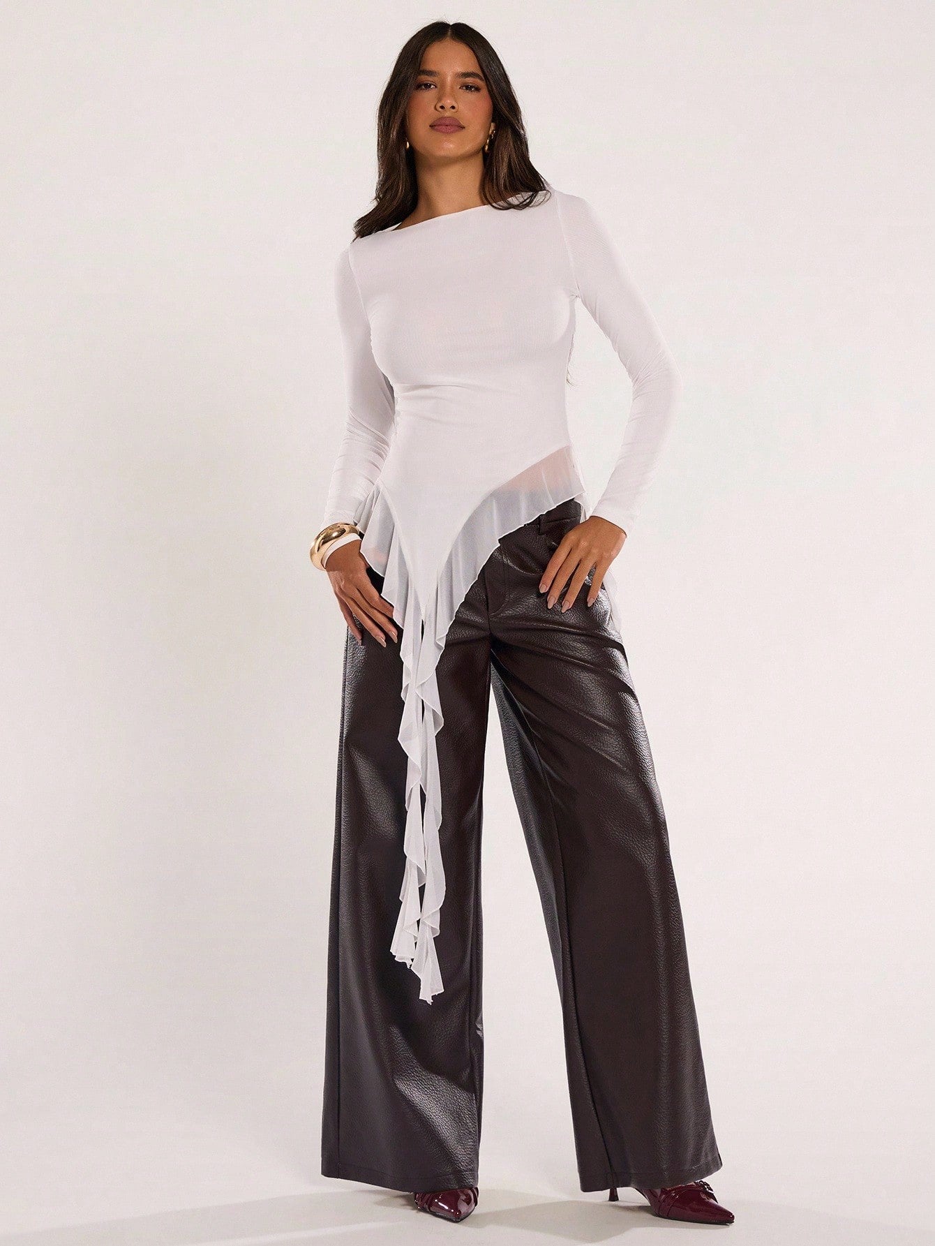 Woman wearing an off-white fitted long sleeve top with an asymmetric ruffle hem paired with dark brown faux leather pants