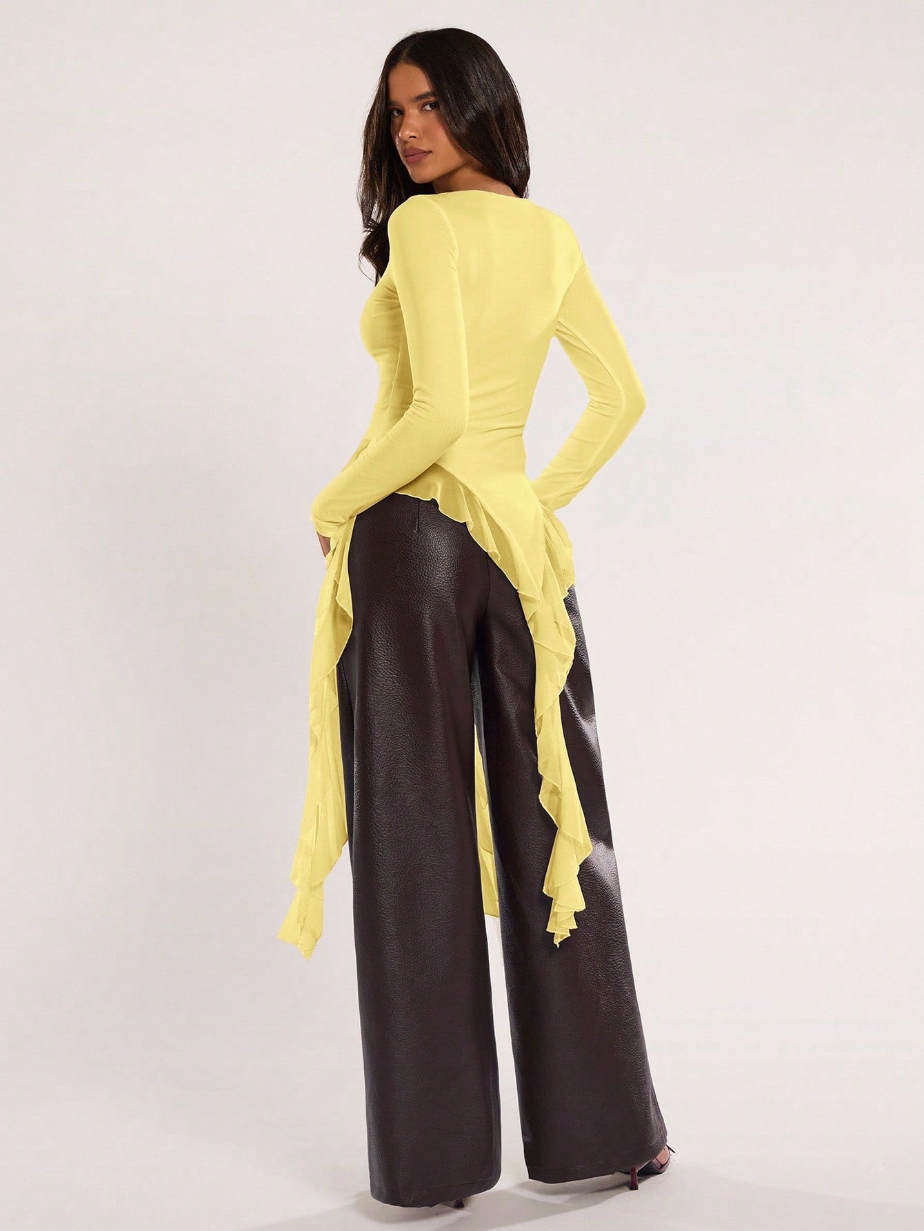 Woman wearing an yellow fitted long sleeve top with an asymmetric ruffle hem paired with dark brown faux leather pants