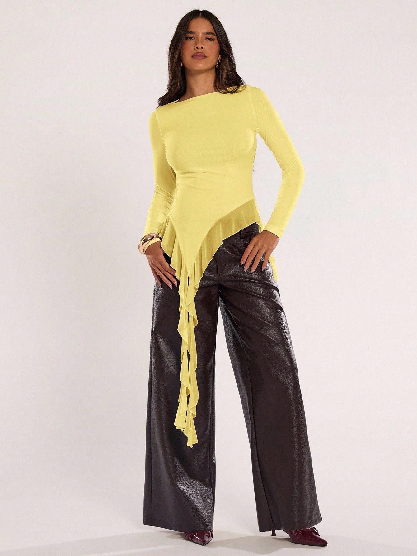 Woman wearing an yellow fitted long sleeve top with an asymmetric ruffle hem paired with dark brown faux leather pants