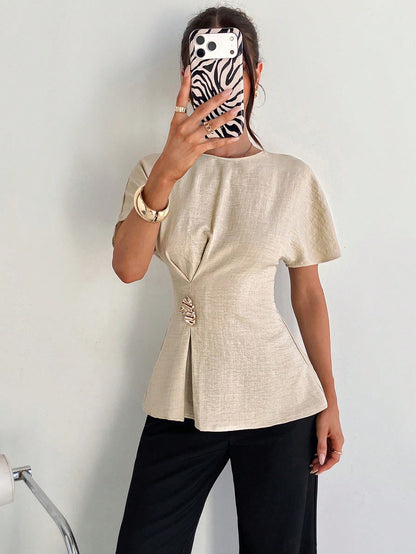 Women's apricot short-sleeve round neck blouse with a statement gold side waist buckle paired with black trousers for an office look.
