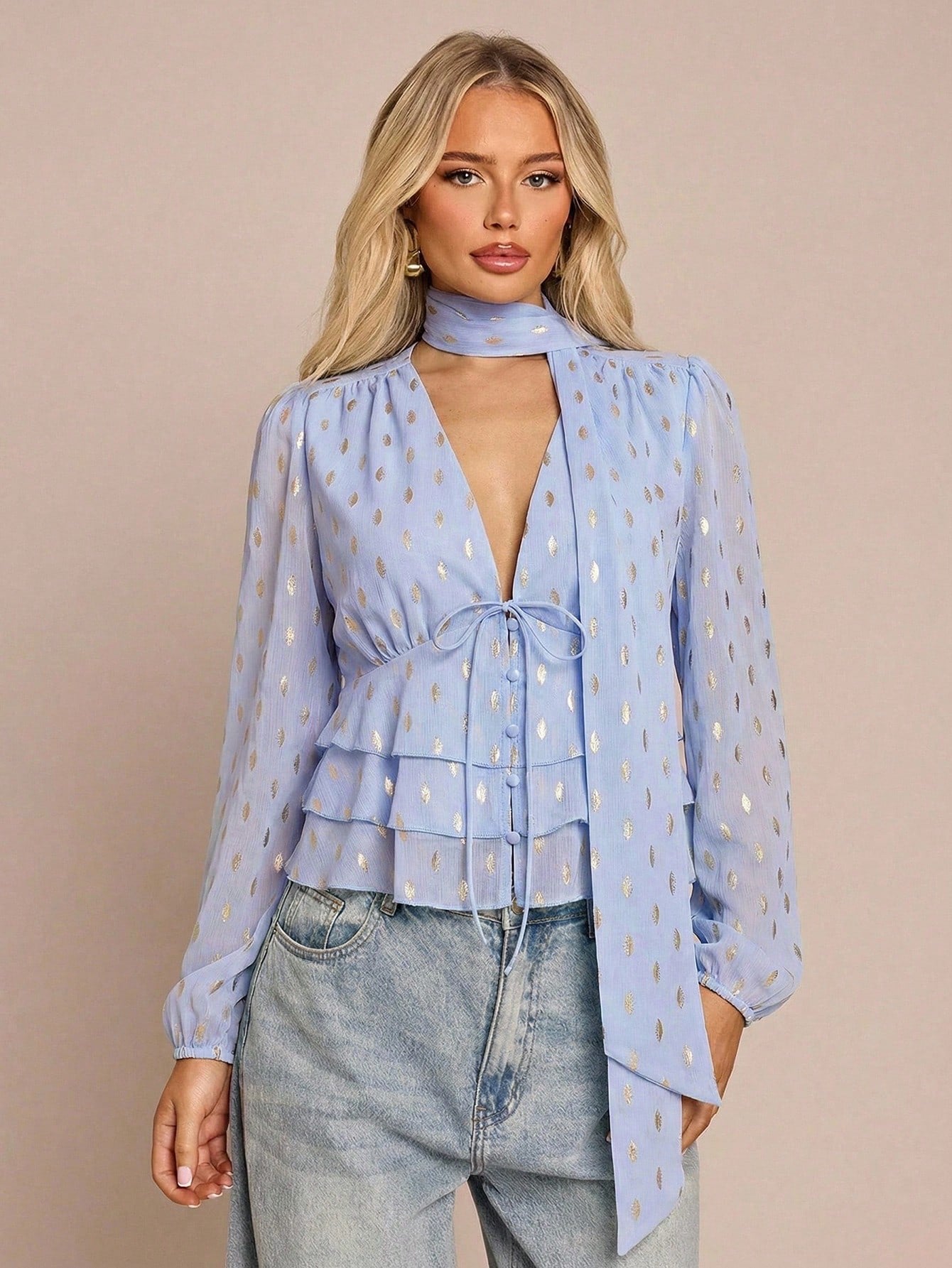 Women's baby blue long-sleeve chiffon blouse with metallic foil dots, a V-neck, a scarf tie, and a tiered ruffled hem paired with blue jeans.