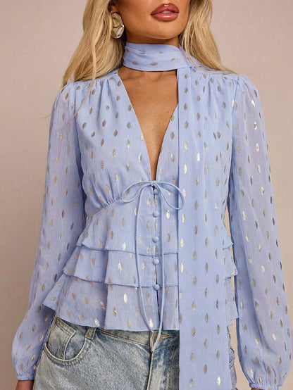 Women's baby blue long-sleeve chiffon blouse with metallic foil dots, a V-neck, a scarf tie, and a tiered ruffled hem paired with blue jeans.