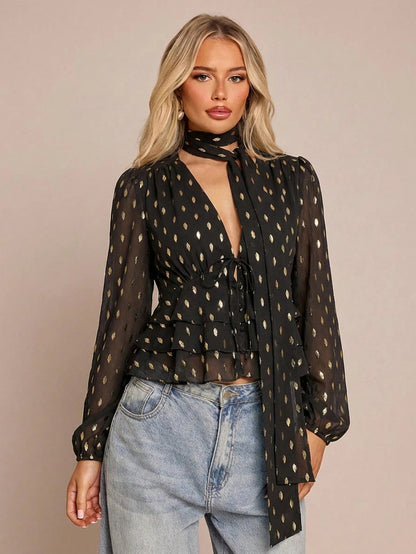 Women's black long-sleeve chiffon blouse with metallic foil dots, a V-neck, a scarf tie, and a tiered ruffled hem paired with blue jeans.