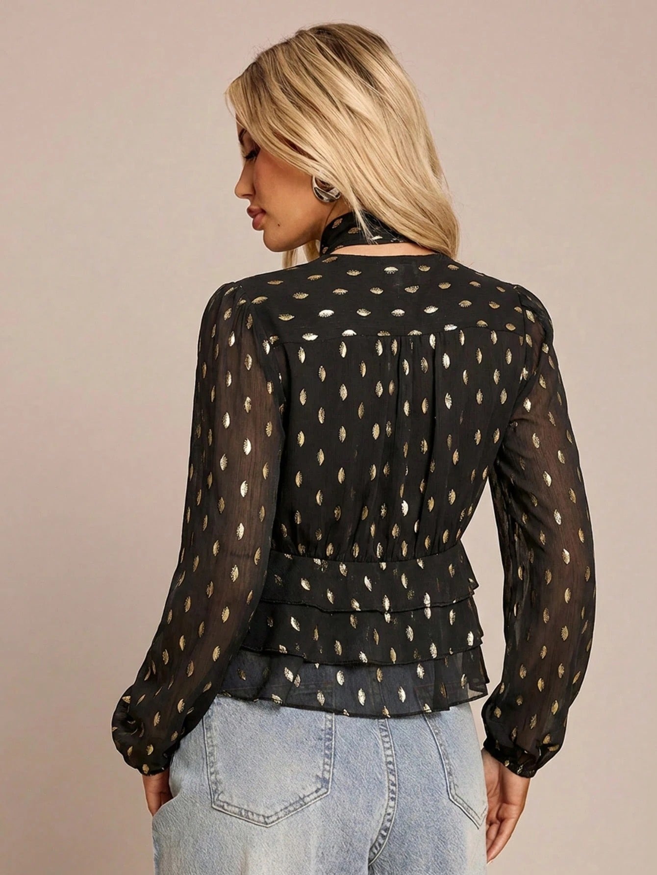 Women's black long-sleeve chiffon blouse with metallic foil dots, a V-neck, a scarf tie, and a tiered ruffled hem paired with blue jeans.