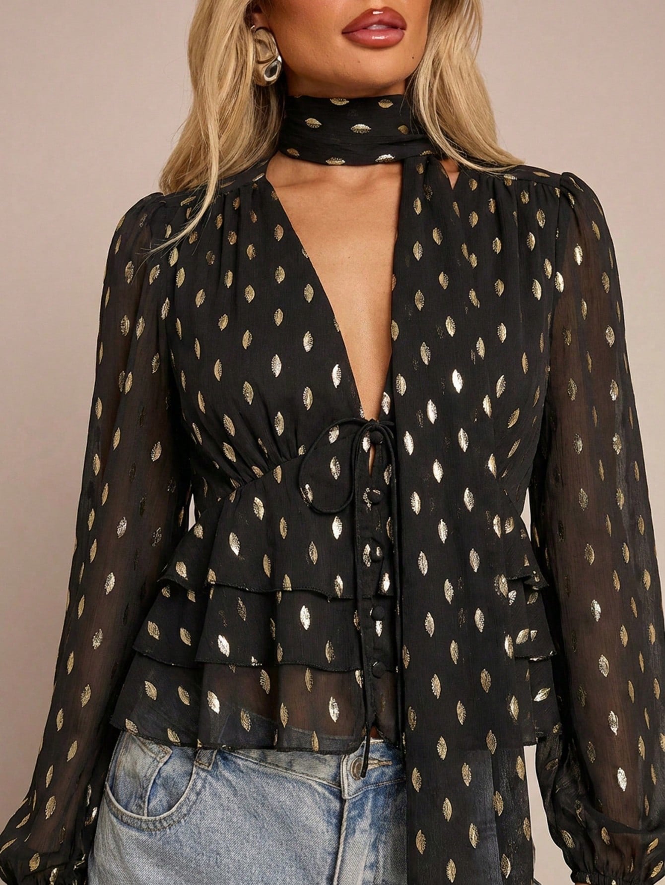 Women's black long-sleeve chiffon blouse with metallic foil dots, a V-neck, a scarf tie, and a tiered ruffled hem paired with blue jeans.