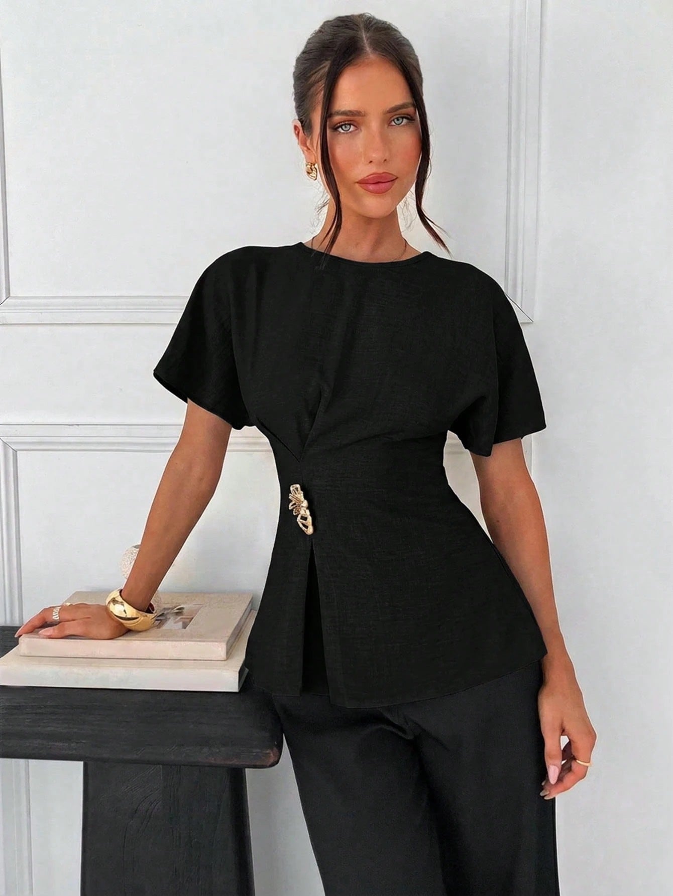 Women's black short-sleeve round neck blouse with a statement gold side waist buckle paired with black trousers for an office look.