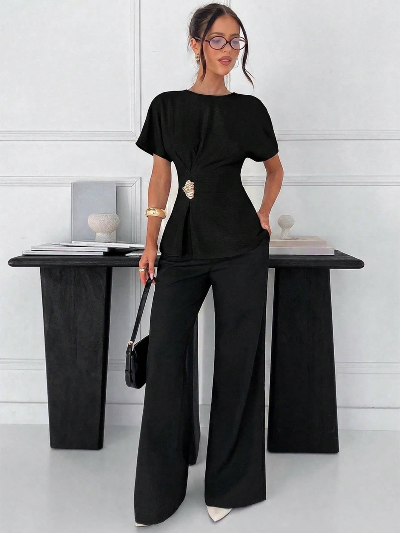 Women's black short-sleeve round neck blouse with a statement gold side waist buckle paired with black trousers for an office look.