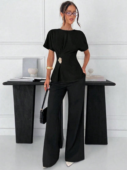 Women's black short-sleeve round neck blouse with a statement gold side waist buckle paired with black trousers for an office look.