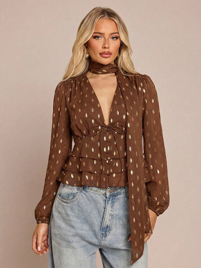 Women's brown long-sleeve chiffon blouse with metallic foil dots, a V-neck, a scarf tie, and a tiered ruffled hem paired with blue jeans.
