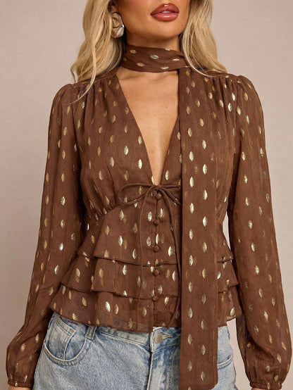 Women's brown long-sleeve chiffon blouse with metallic foil dots, a V-neck, a scarf tie, and a tiered ruffled hem paired with blue jeans.
