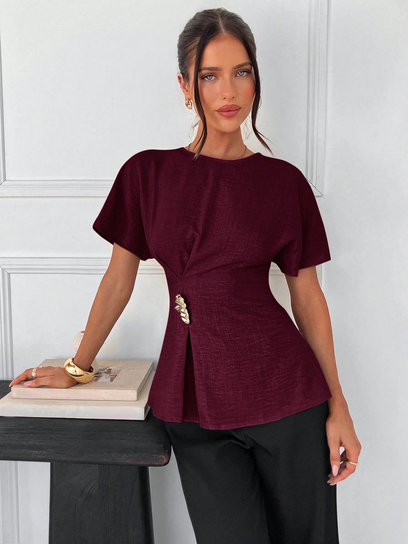 Women's burgundy short-sleeve round neck blouse with a statement gold side waist buckle paired with black trousers for an office look.