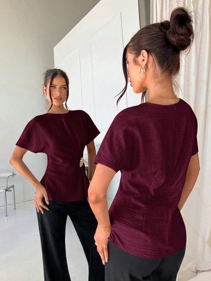 Women's burgundy short-sleeve round neck blouse with a statement gold side waist buckle paired with black trousers for an office look.