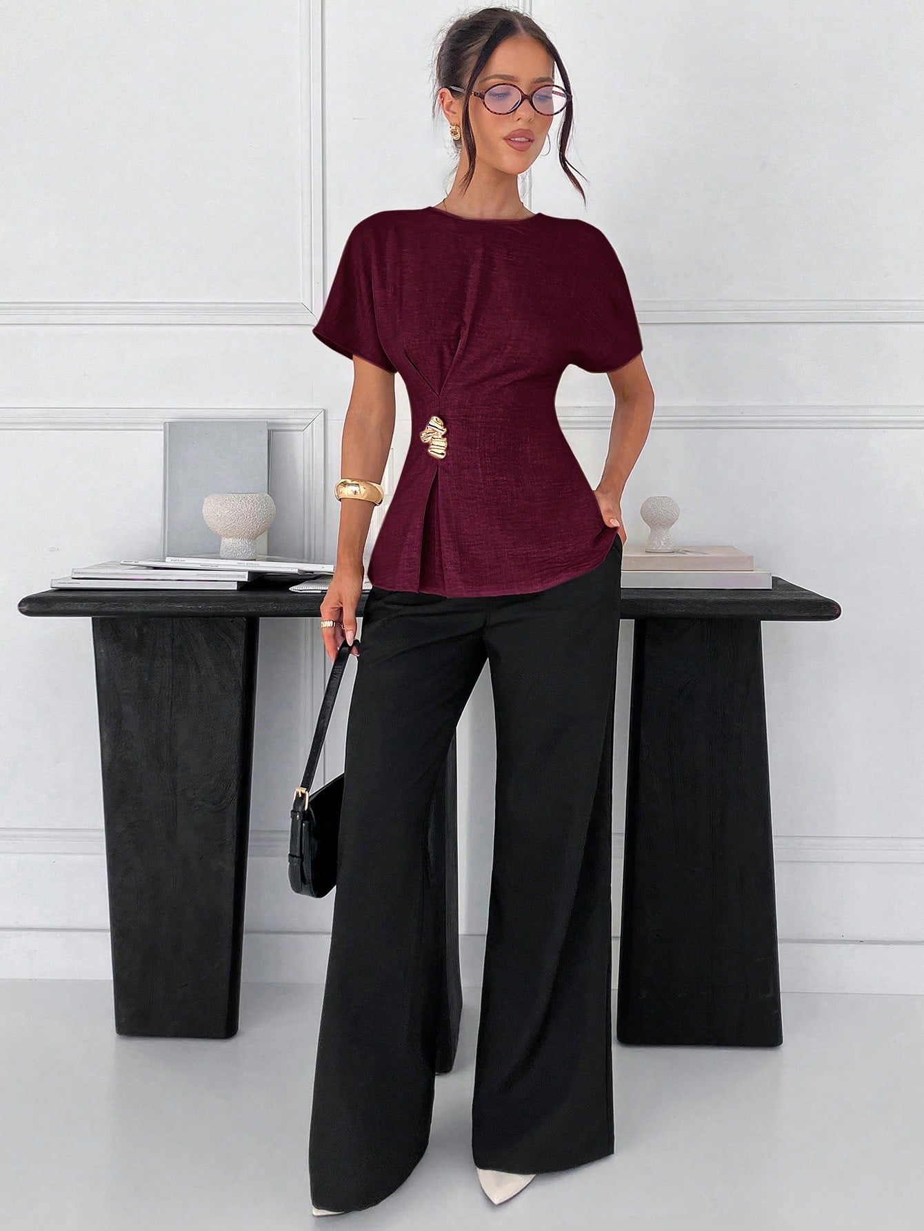 Women's burgundy short-sleeve round neck blouse with a statement gold side waist buckle paired with black trousers for an office look.