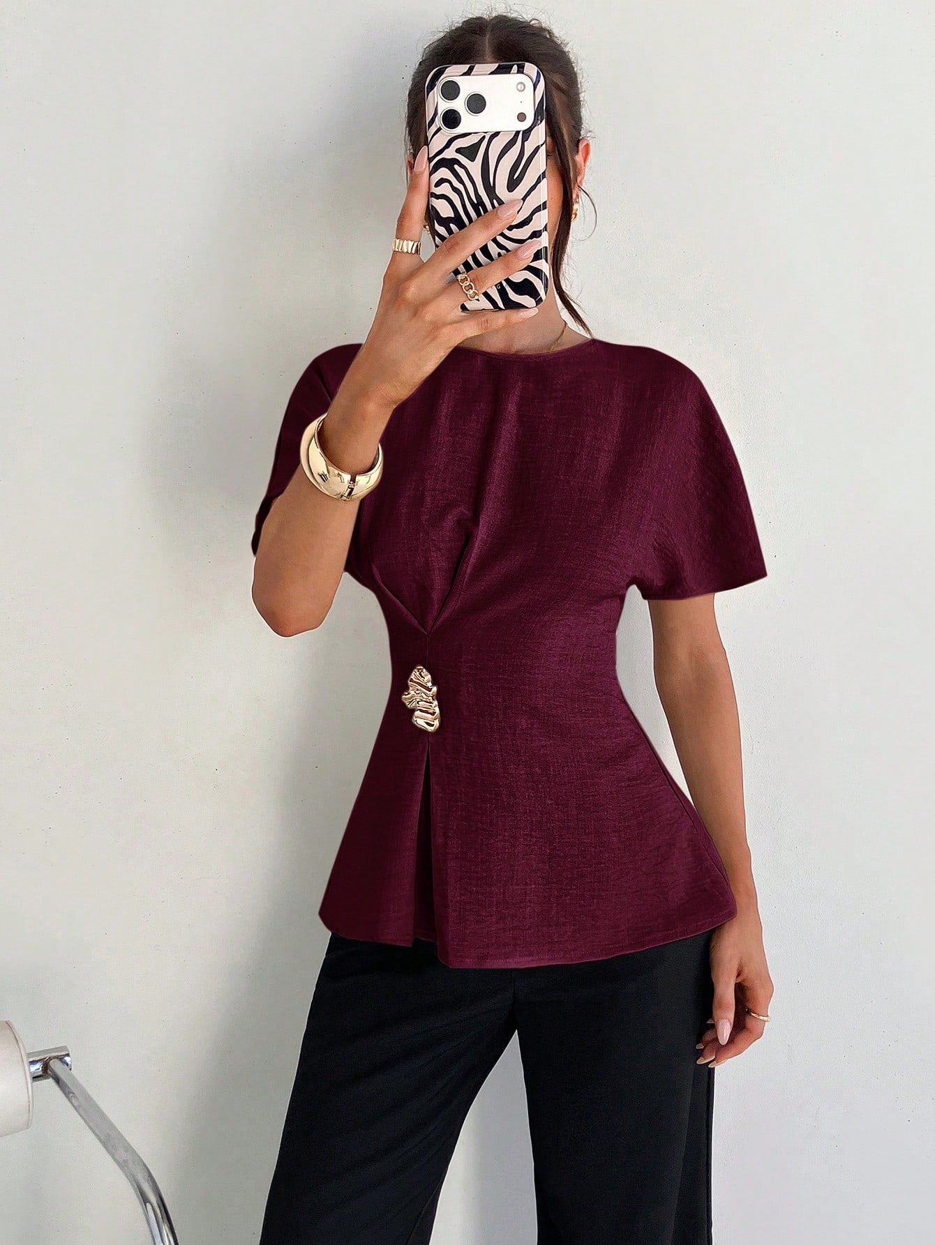Women's burgundy short-sleeve round neck blouse with a statement gold side waist buckle paired with black trousers for an office look.