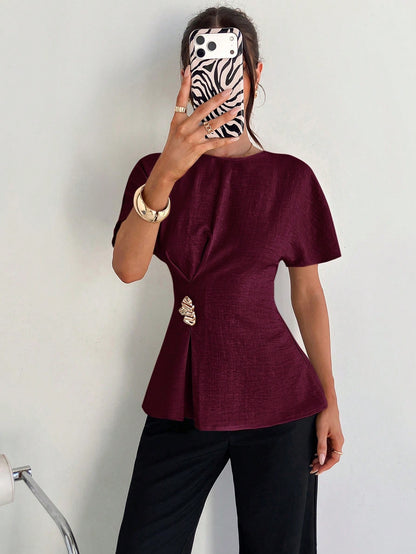 Women's burgundy short-sleeve round neck blouse with a statement gold side waist buckle paired with black trousers for an office look.