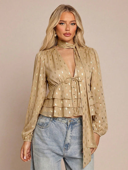Women's khaki long-sleeve chiffon blouse with metallic foil dots, a V-neck, a scarf tie, and a tiered ruffled hem paired with blue jeans.