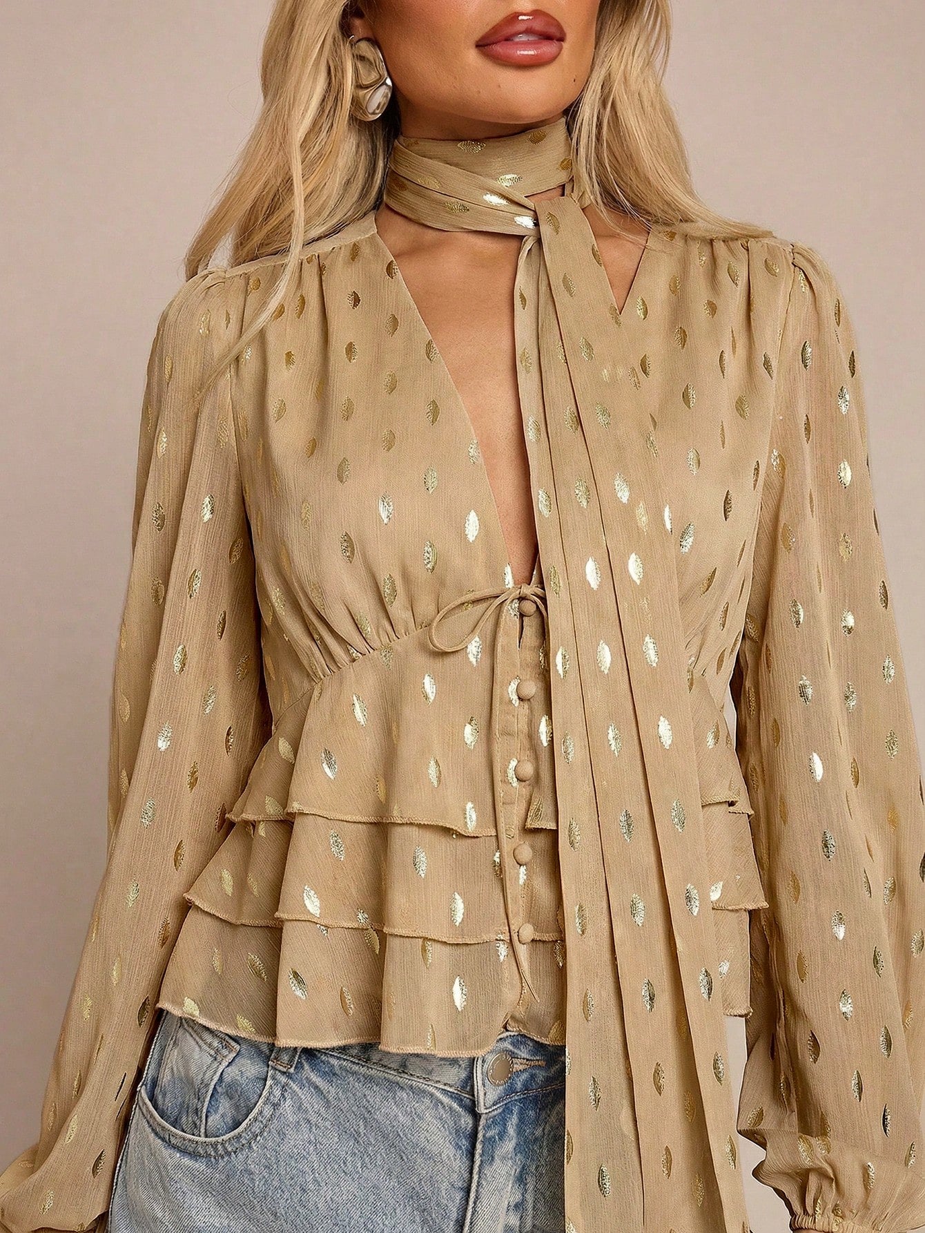 Women's khaki long-sleeve chiffon blouse with metallic foil dots, a V-neck, a scarf tie, and a tiered ruffled hem paired with blue jeans.