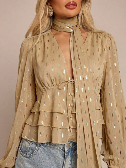 Women's khaki long-sleeve chiffon blouse with metallic foil dots, a V-neck, a scarf tie, and a tiered ruffled hem paired with blue jeans.