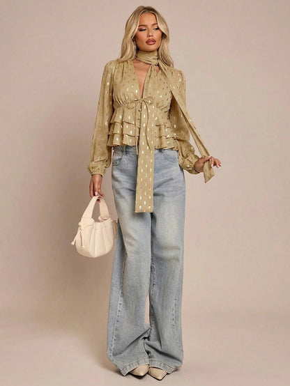 Women's khaki long-sleeve chiffon blouse with metallic foil dots, a V-neck, a scarf tie, and a tiered ruffled hem paired with blue jeans.