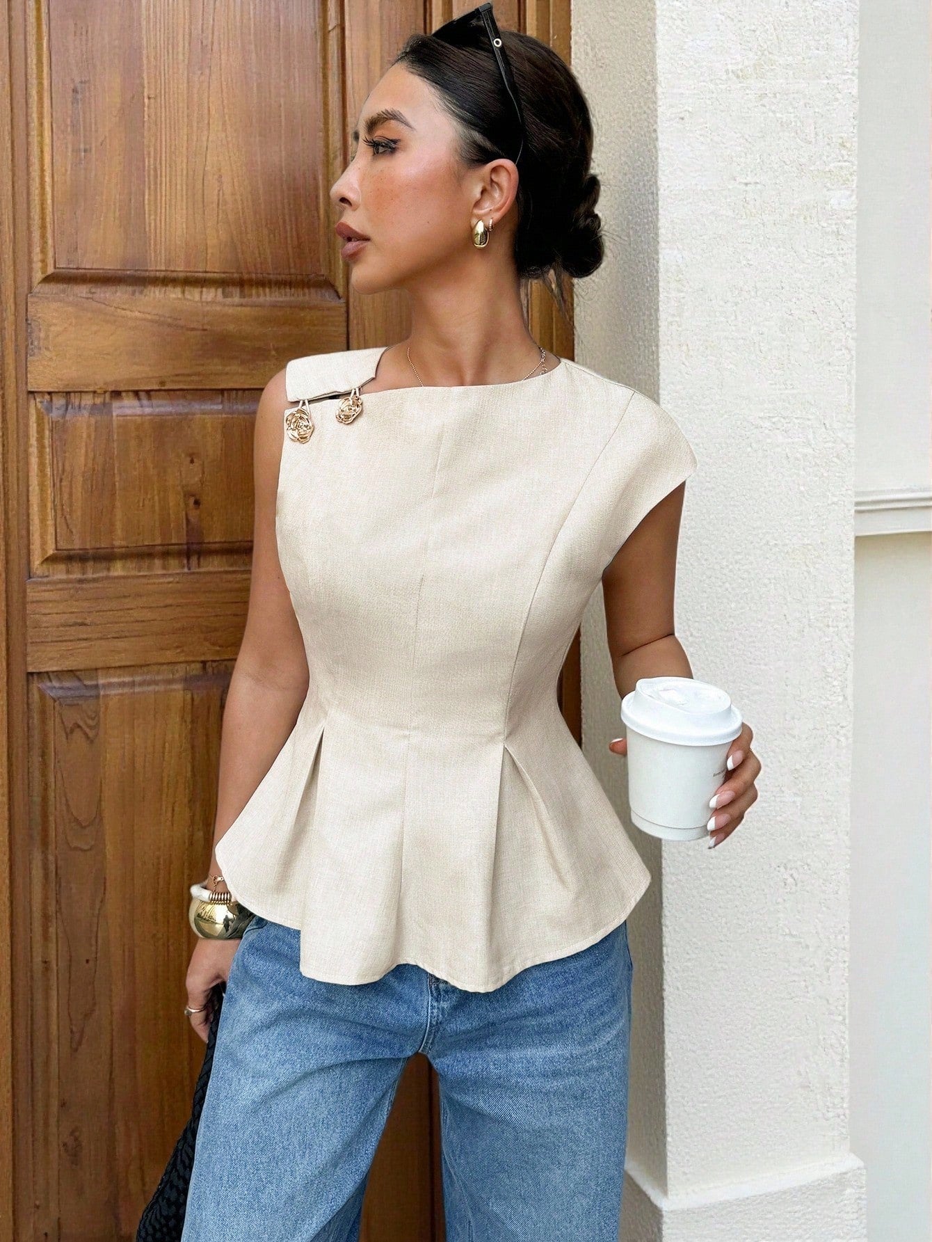 Women's minimalist apricot sleeveless peplum blouse with gold button accents and a pleated flare hem paired with blue jeans.