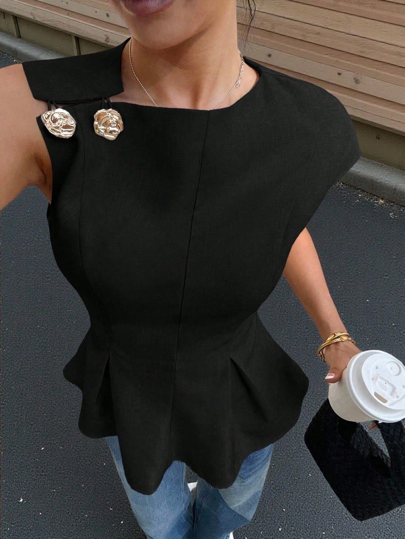 Women's minimalist black sleeveless peplum blouse with gold button accents and a pleated flare hem paired with blue jeans.